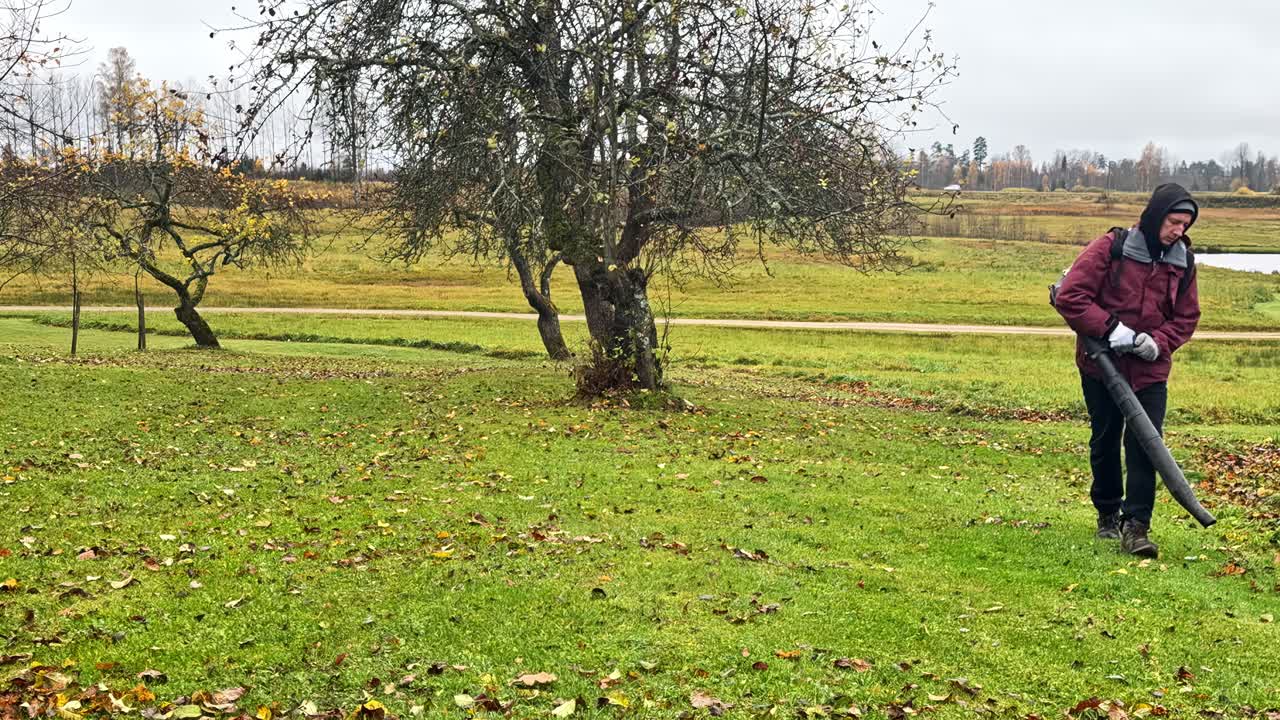 Un uomo usa un soffiatore per foglie per rimuovere i detriti da un prato in una giornata calma, mentre le trame del giardino cambiano con un movimento delicato