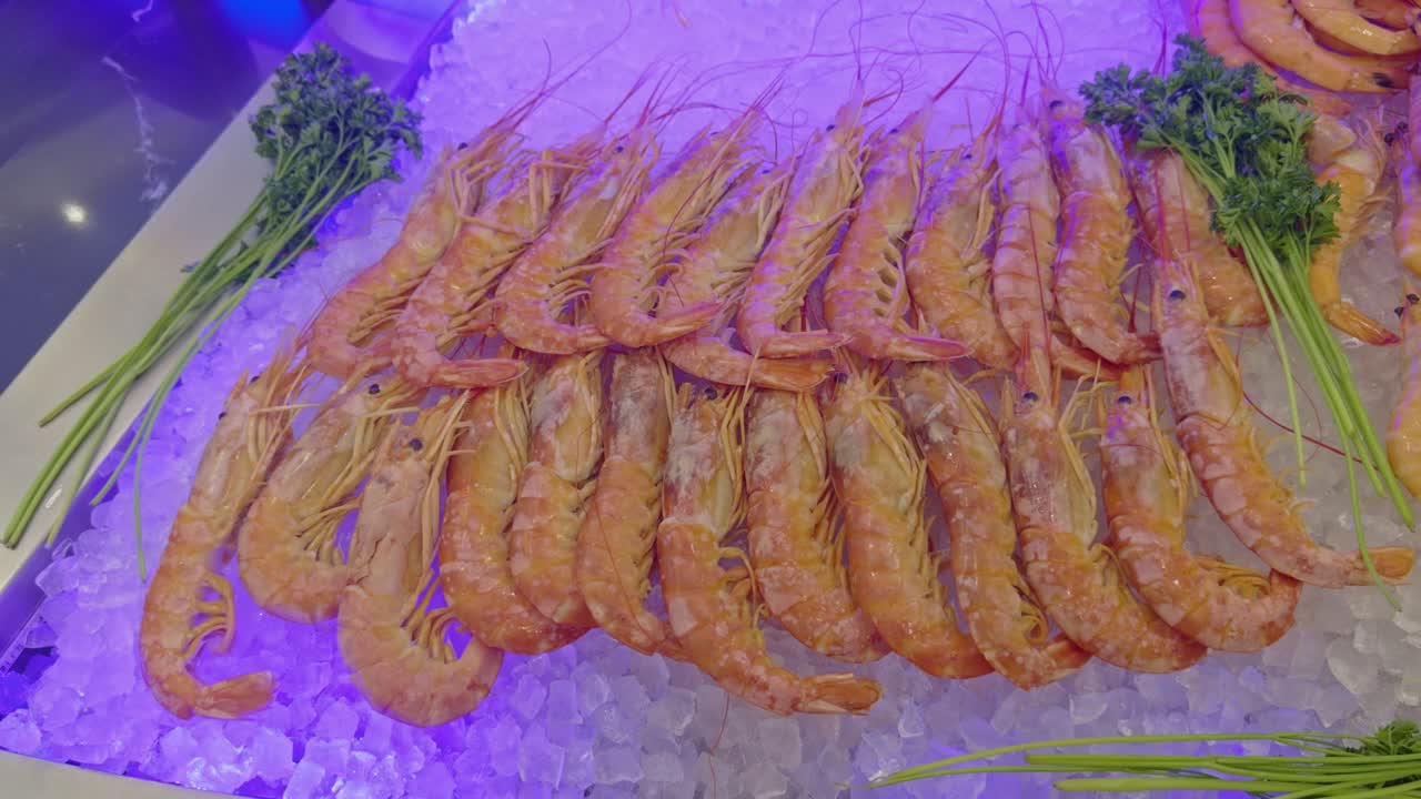 Slow panning shot of fresh langoustines arranged on crushed ice at a self-service seafood buffet. The shellfish are neatly aligned and garnished with green herbs, highlighting freshness and abundance