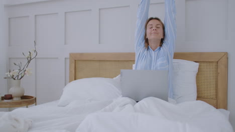 happy woman with laptop lying in bed and smiling. girl in a man's shirt working on a laptop in the early morning