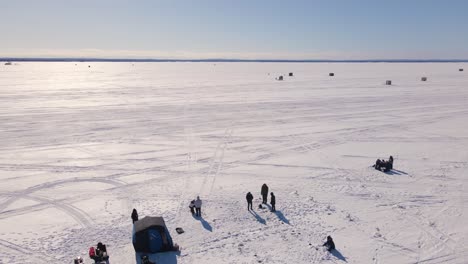 a wide angle drone shot panning upwards showing a beautiful scenery of many people ice fishing during a cold winters day and ice fishing huts in the background in a remote region of canada