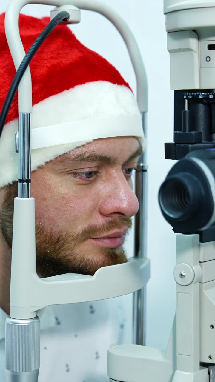 Patient in Christmas cap at ophthalmologist's. Man testing his eyes in clinic. Young male patient checking his eyesight. Vertical video