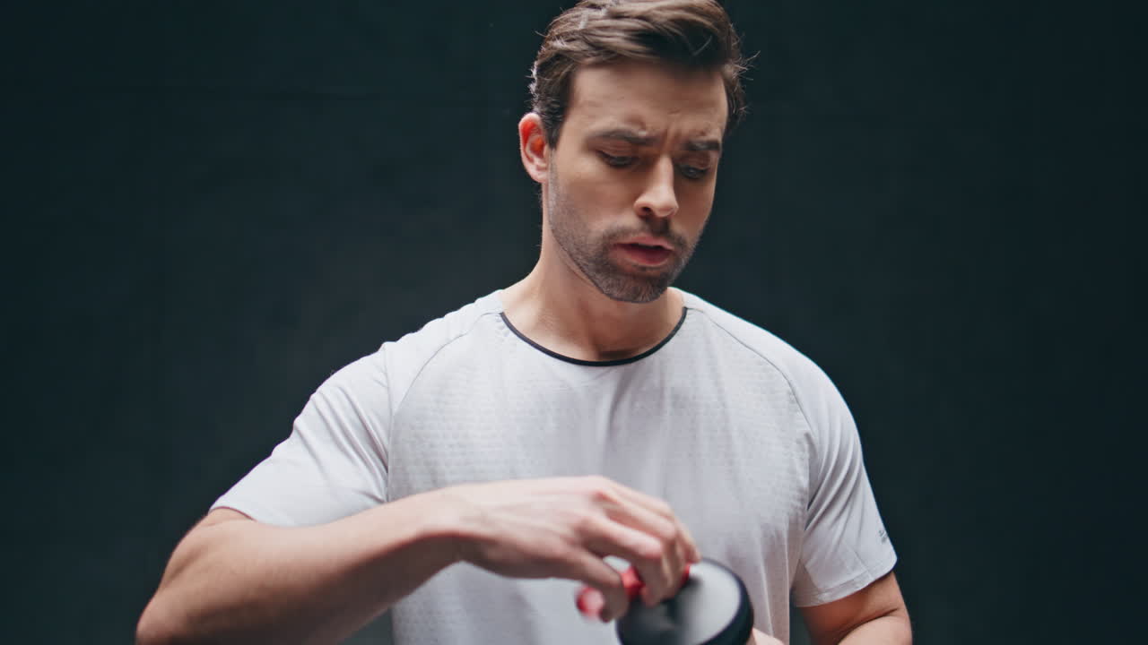 Tired athlete shaking isotonic drink in cup in black wall background closeup