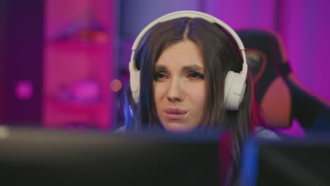 Young woman wearing white headphones stares at screen with disappointed expression after losing game, seated in colorful neon-lit studio with pink and purple lights reflecting mood and emotion