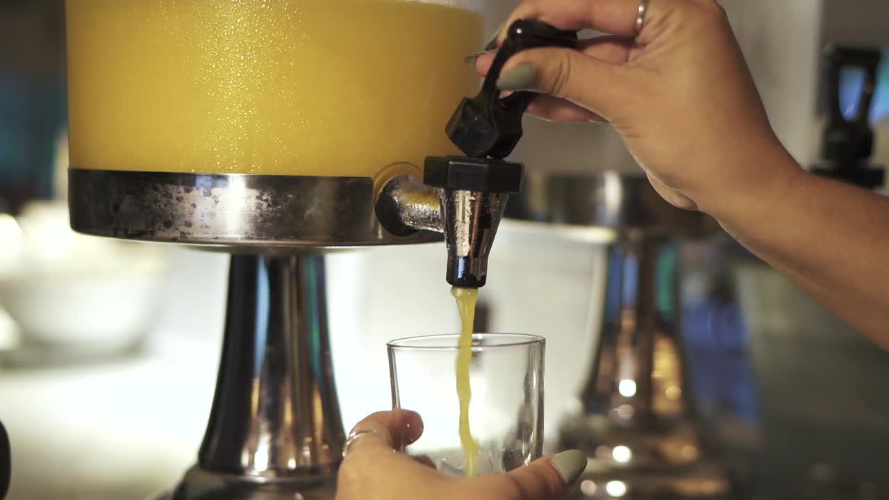 fotografía de primer plano de un huésped de un hotel que sirve jugo de naranja de un dispensador en el comedor durante el desayuno