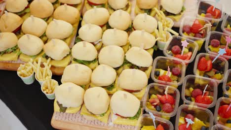 a beautifully arranged catering buffet table offers a delicious variety of fresh finger foods, including mini burgers, croissant sandwiches, fruit cups, and canapés for a party or event.