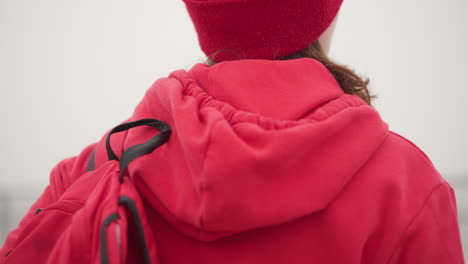 vista de cerca de la mujer con capucha roja y gorra ajustando la correa de la bolsa roja en un entorno exterior brumoso, creando una acogedora vibra de invierno con un fondo borroso de un paisaje atmosférico sereno