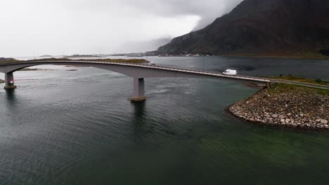 A-white-camper-van-crossing-a-bridge-over-a-Fjord-on-a-moody-day,-the-clouds-sitting-over-the-majestic-mountain-coastline,-Lofoten-Islands,-Norway