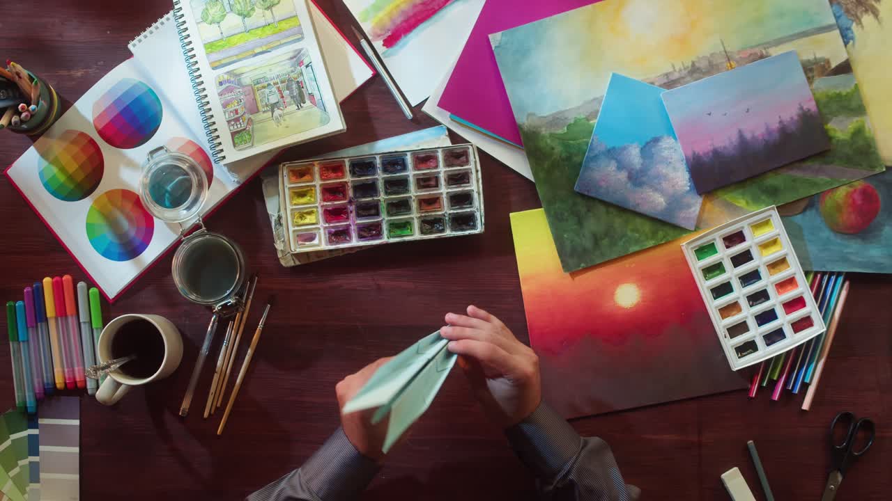 Hands Folding Paper Airplane on Artist's Desk with Art Supplies