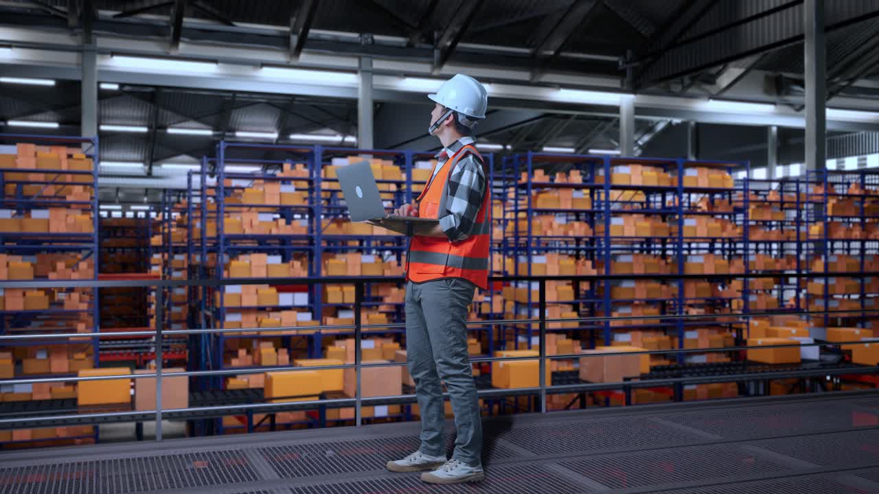 vista lateral completa del cuerpo de un ingeniero masculino asiático con casco de seguridad mirando una computadora portátil y mirando a su alrededor mientras está de pie en el almacén con estantes llenos de productos de entrega