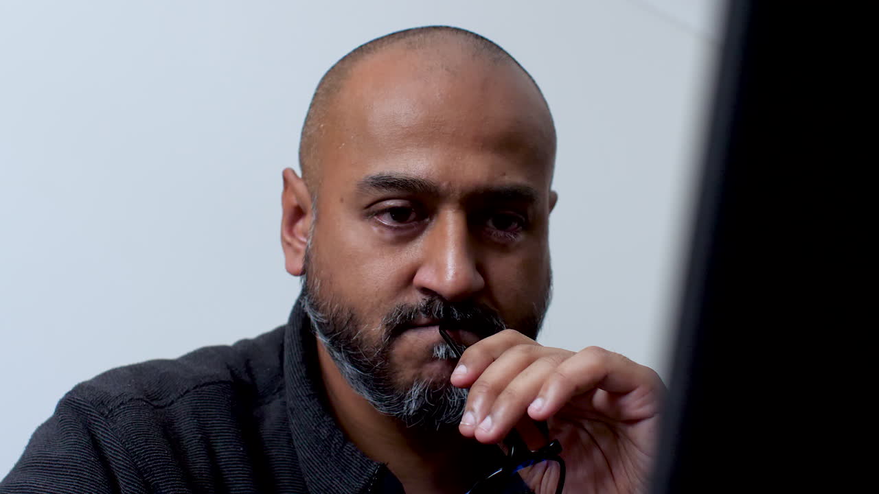 Bald male feeling anxious, holding glasses in his mouth, and working on a laptop