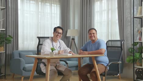médico asiático con un paciente gordo en la clínica, escribiendo recetas, sentado en el escritorio y sonriendo a la cámara.