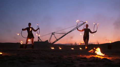 a group of men and woman fire show at night on the sand against the background of fire and tower cranes.