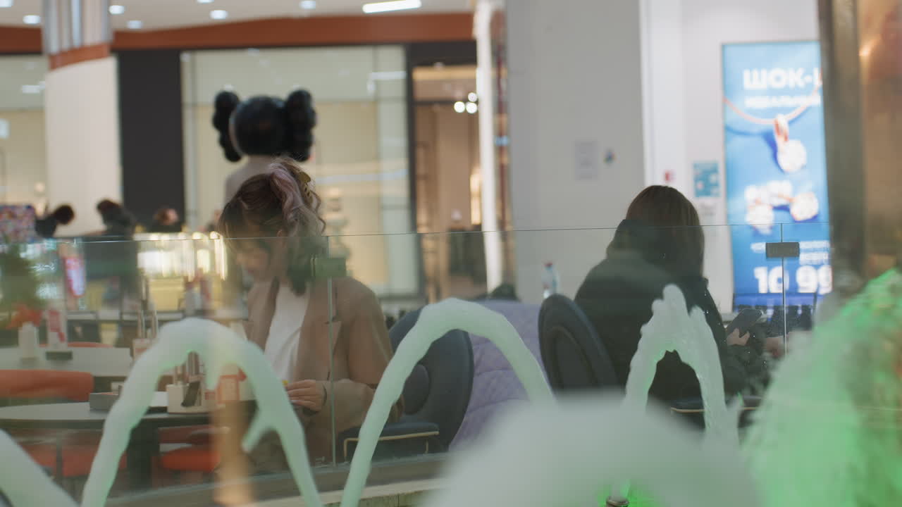 전경에 흰색 분수 모양이 있고, 캐주얼한 좌석 공간과 소매점 간판이 보이는 유리창 너머로 쇼핑객을 넓게 촬영했으며, 부드러운 반사와 주변 조명이 있는 자연스러운 도시 여가 장면입니다.