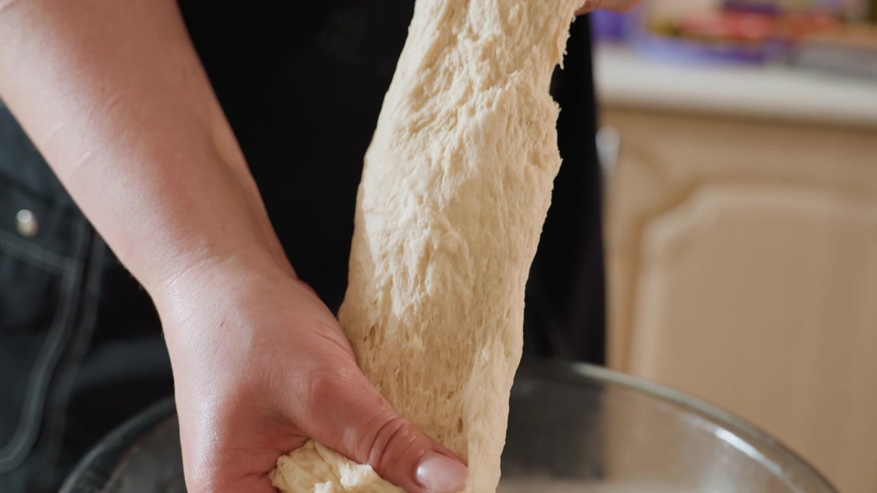 Vista cercana muestra manos presionando, doblando y estirando masa cruda elástica dentro de un bol de cristal para mezclar sobre la encimera de la cocina, sugiriendo la preparación de pan casero con movimiento táctil y luz natural.