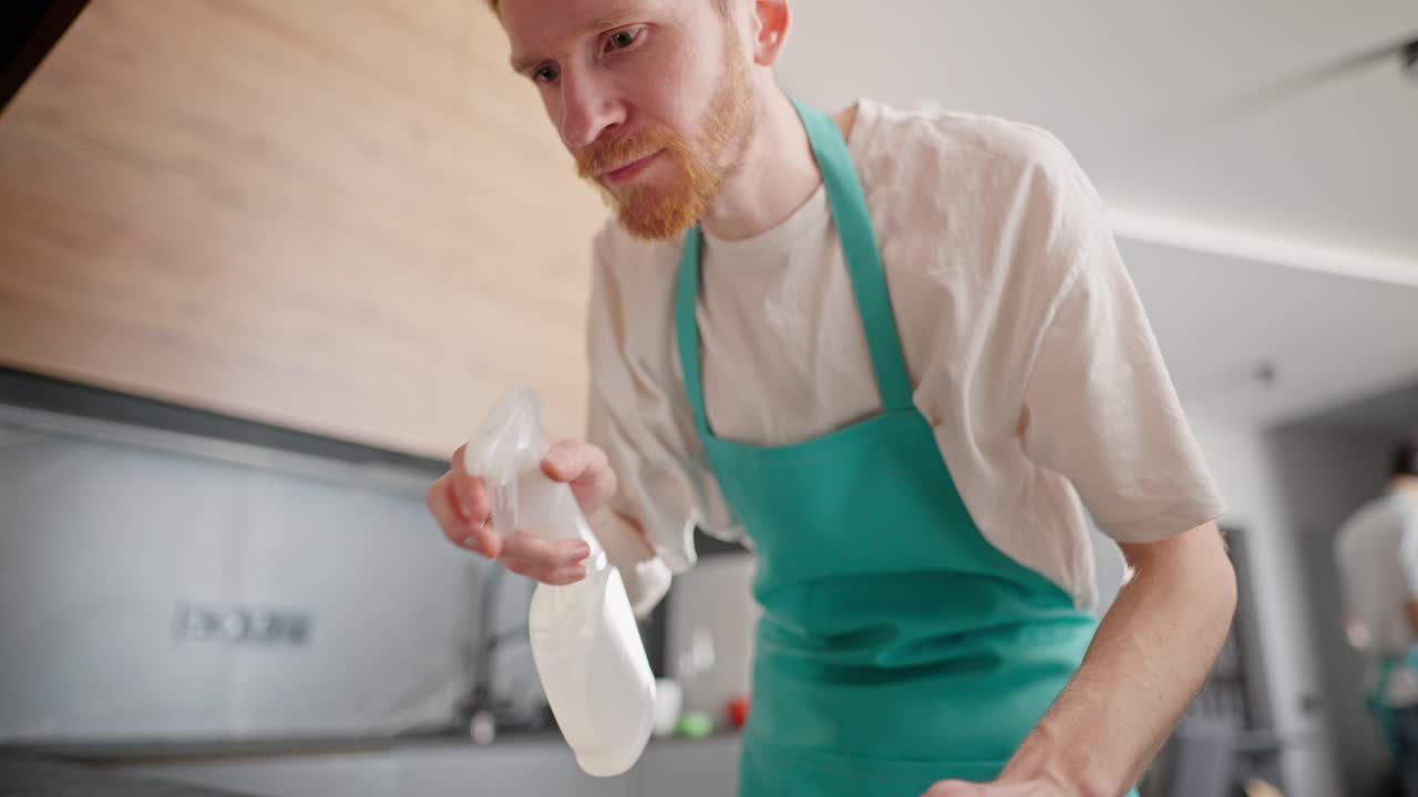Camera view of a blond guy in a white T-shirt and a blue apron as a cleaner washes the table in the kitchen in a modern apartment. Cleaning company in the apartment on call
