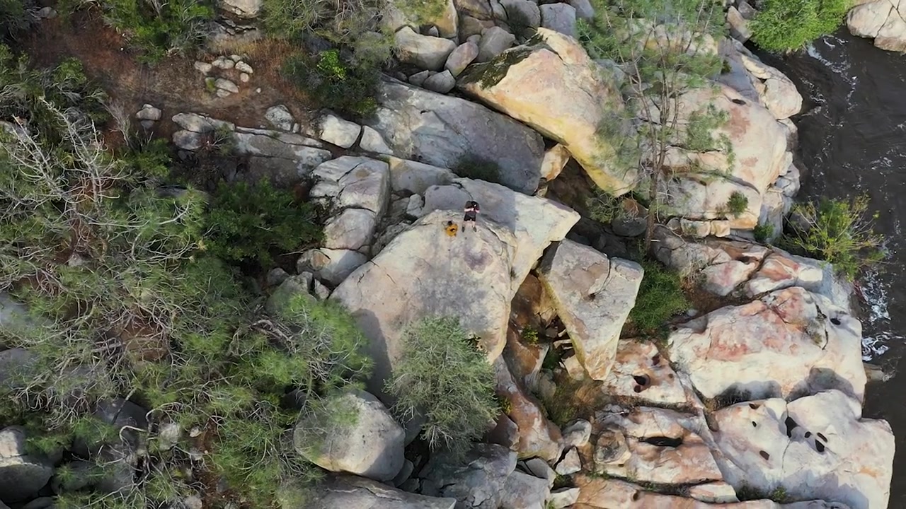young Caucasian male hiker in a black tee shirt taking photos with a camera on top of a large boulder on the kern valley river in California during sunset aerial orbit