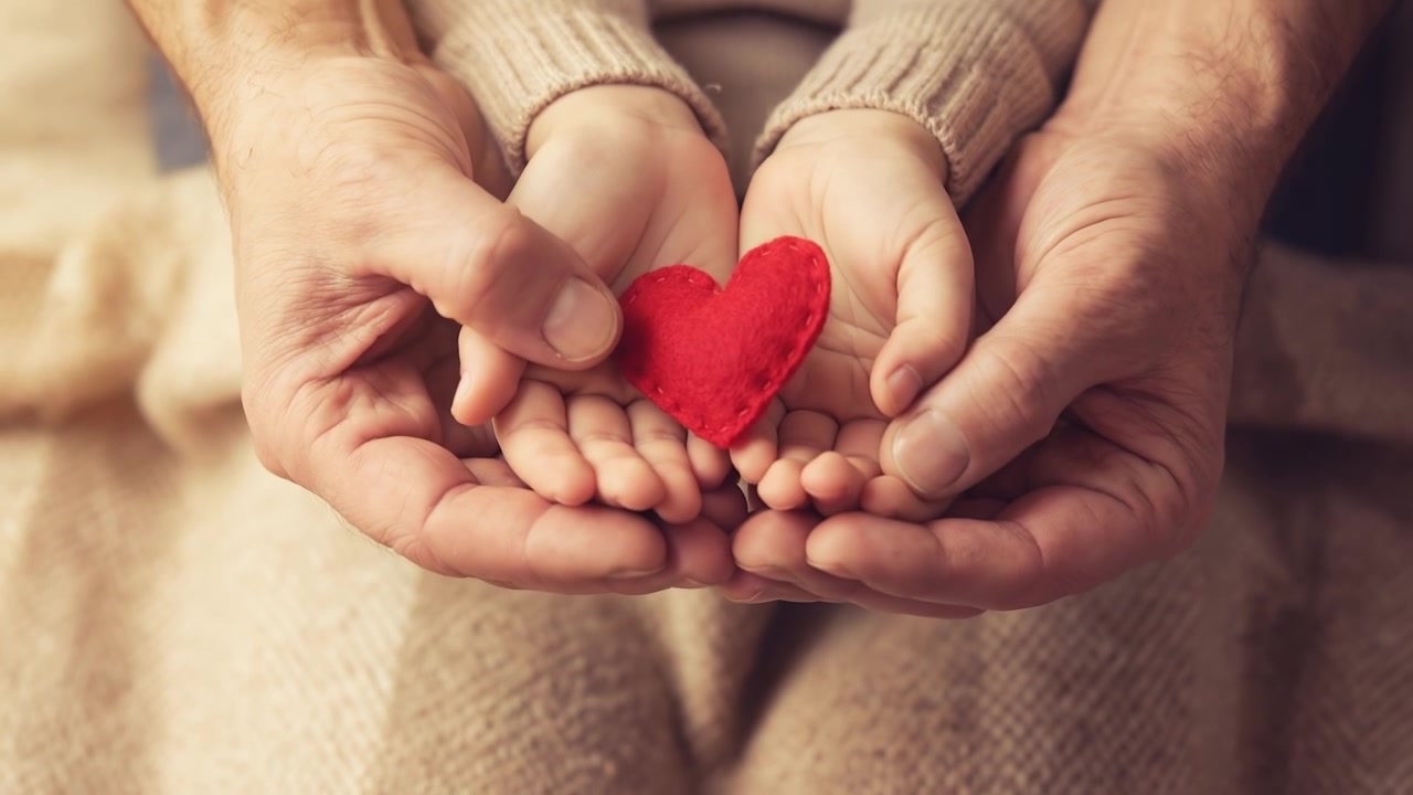 Manos de adulto y niño sosteniendo juntos un pequeño corazón rojo sobre una cálida manta de punto.