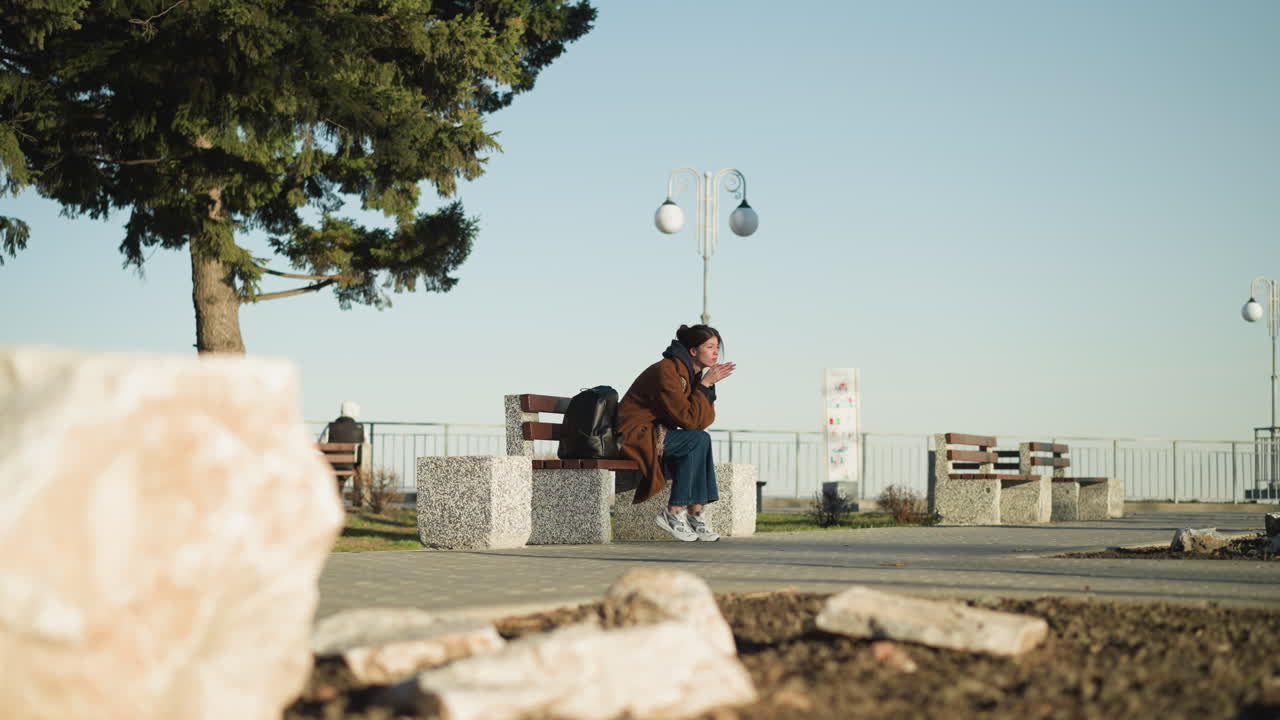 una mujer joven con un abrigo marrón, pantalones vaqueros azules y zapatos blancos se sienta sola en un banco en un parque urbano, su mochila negra a su lado. parece deprimida, con el rostro apoyado en sus manos