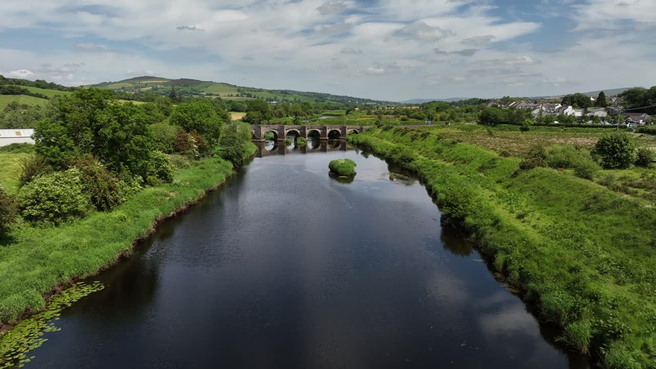 Clady Bridge, County Tyrone, Northern Ireland, June 2023. Drone approach along River Finn to arched bridge with sloped hills in backdrop along grassy riverbanks.