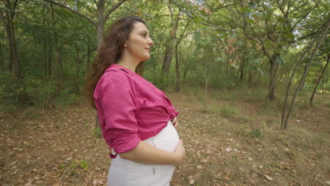 tracking shot of pregnant woman expectant mother walking alone in forest, attractive caucasian casual dressed mom stroking caress touching her belly with hands and smiling, childbearing and tenderness