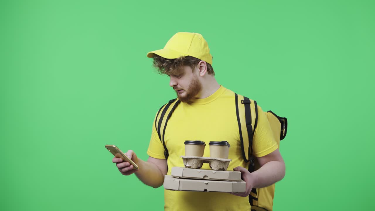 Portrait of men holding coffee and pizza, specifying delivery address using smartphone. Male in yellow courier uniform with thermo bag posing on green screen. Close up. Slow motion ready 59.97fps