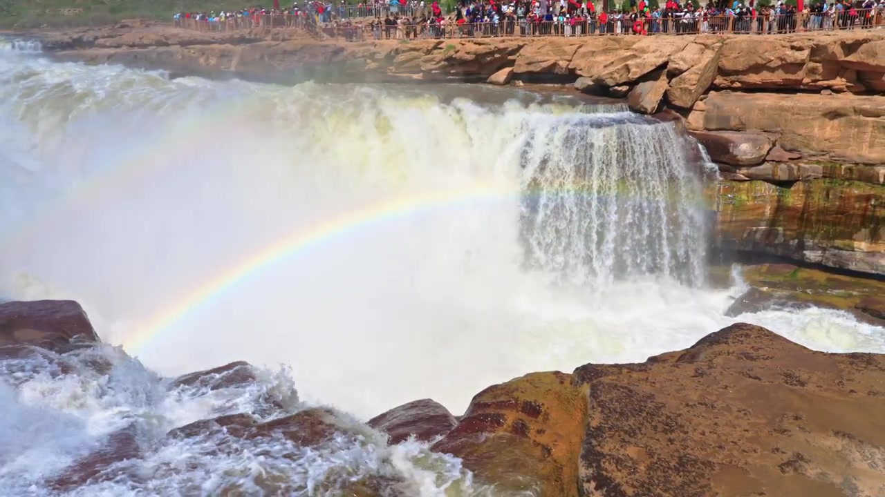 Detaillierte Draufsicht auf den Scheitelpunkt des Hukou-Wasserfalls. Ein lebhafter doppelter Regenbogen spannt sich über das aufgewühlte weiße Wasser und den aufsteigenden Nebel, während der Gelbe Fluss vorwärts strömt.