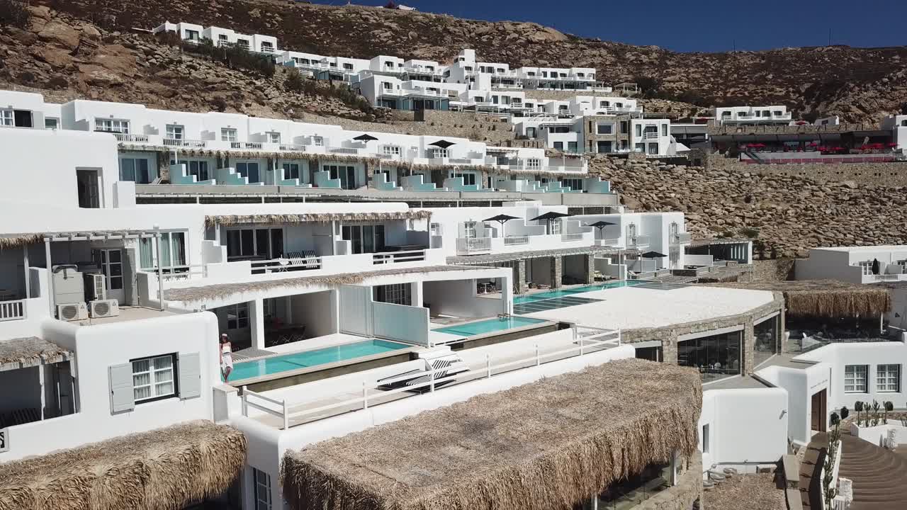 mujer vestida de verano caminando por la piscina privada de la villa con fachada blanca, isla de mykonos, grecia, vista aérea trasera