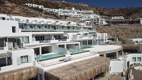 Female-in-Summer-Dress-Walking-by-Private-Villa-Pool-With-White-Facade,-Mykonos-Island,-Greece,-Pull-Back-Aerial-View