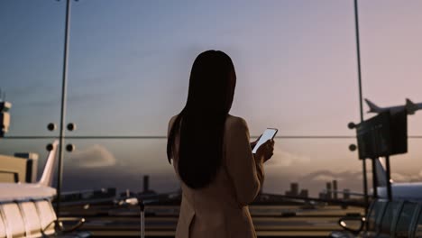 back view of asian businesswoman with rolling suitcase in boarding lounge of airline hub, using smartphone while waiting for flight, airport terminal with airplane takes off outside the window