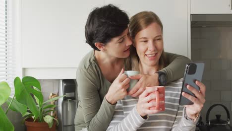 Kaukasische-Frau-Küsst-Ihre-Frau,-Während-Sie-Zu-Hause-In-Der-Küche-Einen-Videoanruf-Auf-Dem-Smartphone-Führt