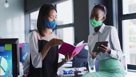 diverse female office colleagues wearing face masks discussing at modern office