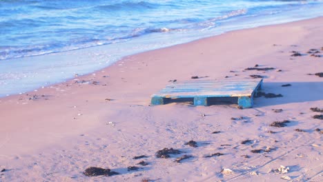 Old-wooden-palette-on-the-shore,-trash-and-waste-litter-on-an-empty-Baltic-sea-white-sand-beach,-environmental-pollution-problem,-golden-hour-light-on-evening,-handheld-medium-shot