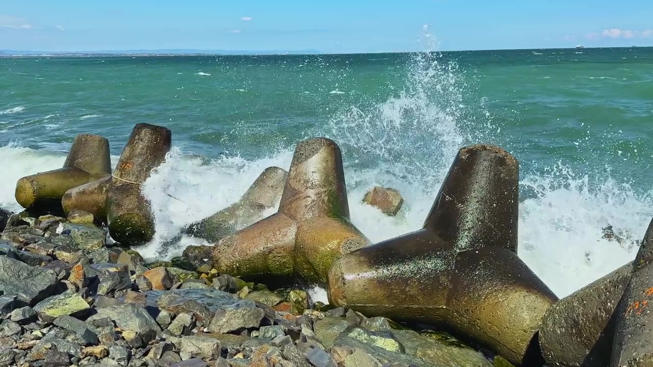 Bølger slår mod betonbarrierer ved kysten på en solrig dag ved havet.