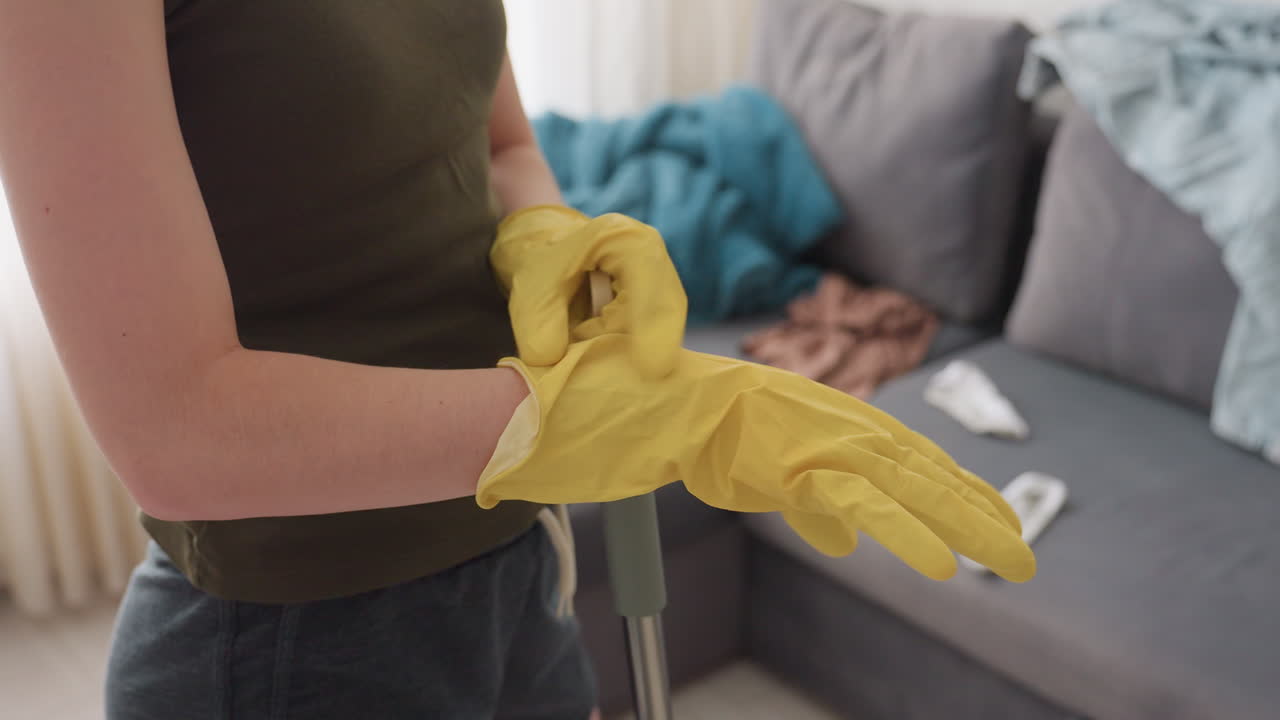 Close up side view of lady adjusting yellow gloves while holding mop in living room near couch with scattered clothes showing preparation for cleaning and focus on household chores