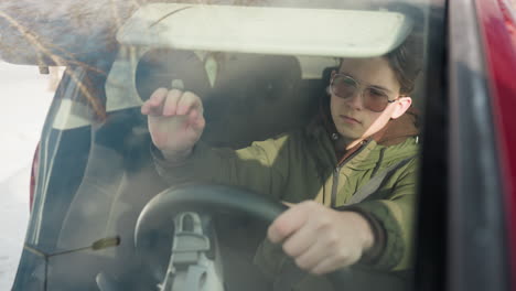 man in green winter jacket and hoodie lowers sun visor while sitting in car with sunlight reflecting off face, wearing sunglasses, looking up with serious expression, seen from windshield angle