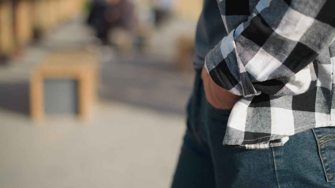 Close up side view of white person strolling slowly with hands in jean pockets on sunlit paved walkway with blurred greenery and urban elements in background