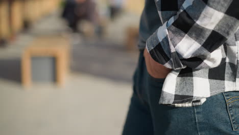 close up side view of white person strolling slowly with hands in jean pockets on sunlit paved walkway with blurred greenery and urban elements in background