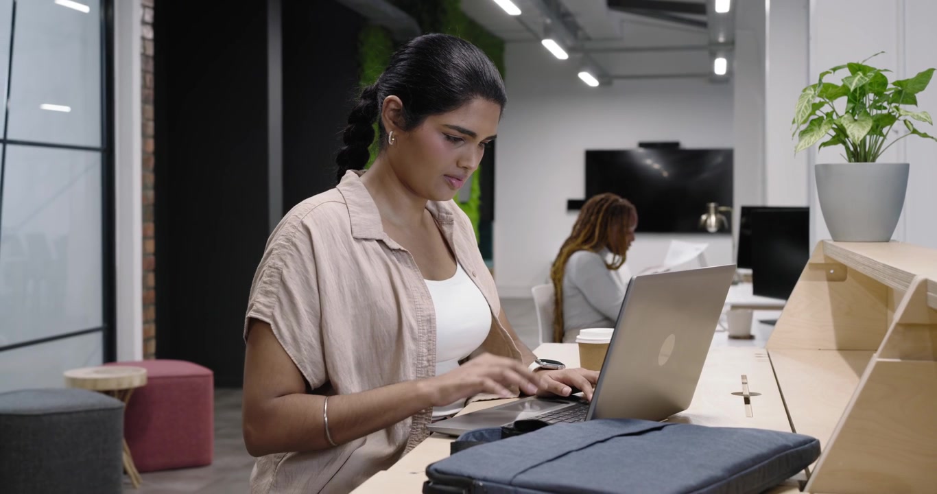 Compañeros de trabajo diversos en el mostrador. Mujer india escribiendo y guardando un portátil plateado en una bolsa azul marino para llevar.