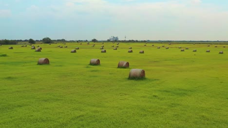 Drone-over-hay-bails-in-field,-factory-in-BG