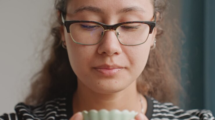 Ospite dell'hotel che inala l'aroma della tazza, rilassato. Luce soffusa dalla finestra, sguardo fisso verso l'alto, riflesso degli occhiali, sorriso sottile, routine mattutina in una suite d'hotel, respiro lento, che pianifica la giornata con calma fiducia.