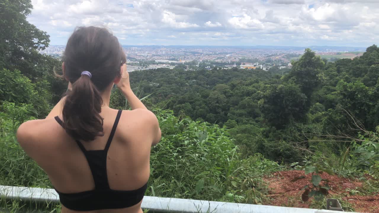 vista trasera de una turista-excursionista tomando una foto panorámica del paisaje urbano usando su teléfono inteligente en el bosque de montaña en un día soleado en praia porto, brasil - toma de primer plano medio