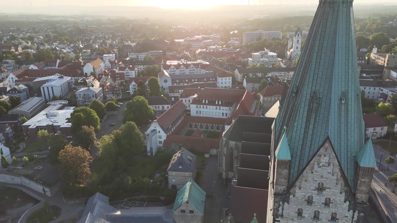 Sunlit drone orbit with warm lens flares frames the cathedral tower beside the General Vicariate administration building and surrounding city.