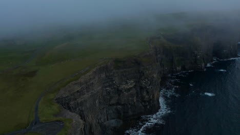 Aerial-shot-of-high-vertical-rock-walls-above-rippled-sea-surface-after-sunset.-View-through-sparse-clouds.-Cliffs-of-Moher,-Ireland