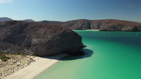 cinematic drone shot of balandra beach, view of red hills, turquoise waters, white-sand beaches, and mountains, revealing beaches past a large bolder