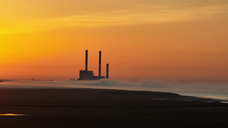 a long focal range drone shot, in lateral motion or side tracking, of a french coal power plant factory shut down , near the loire river, with mist and haze and fog, at sunrise