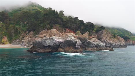 mcway falls shrouded in fog, revealed from behind rocks over the ocean, low aerial