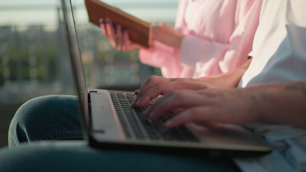 야외에서 노트북 키보드에서 타이핑하는 손의 클로즈업, 다른 사람이 뒷면에 책을 들고 있는 모습, 흐릿한 배경은 초록색과 빛이 특징입니다.