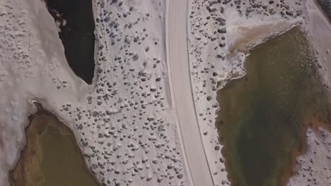 Birdeye-perspective-on-sandy-desert-road-surrounded-by-small-lagoons,-aerial