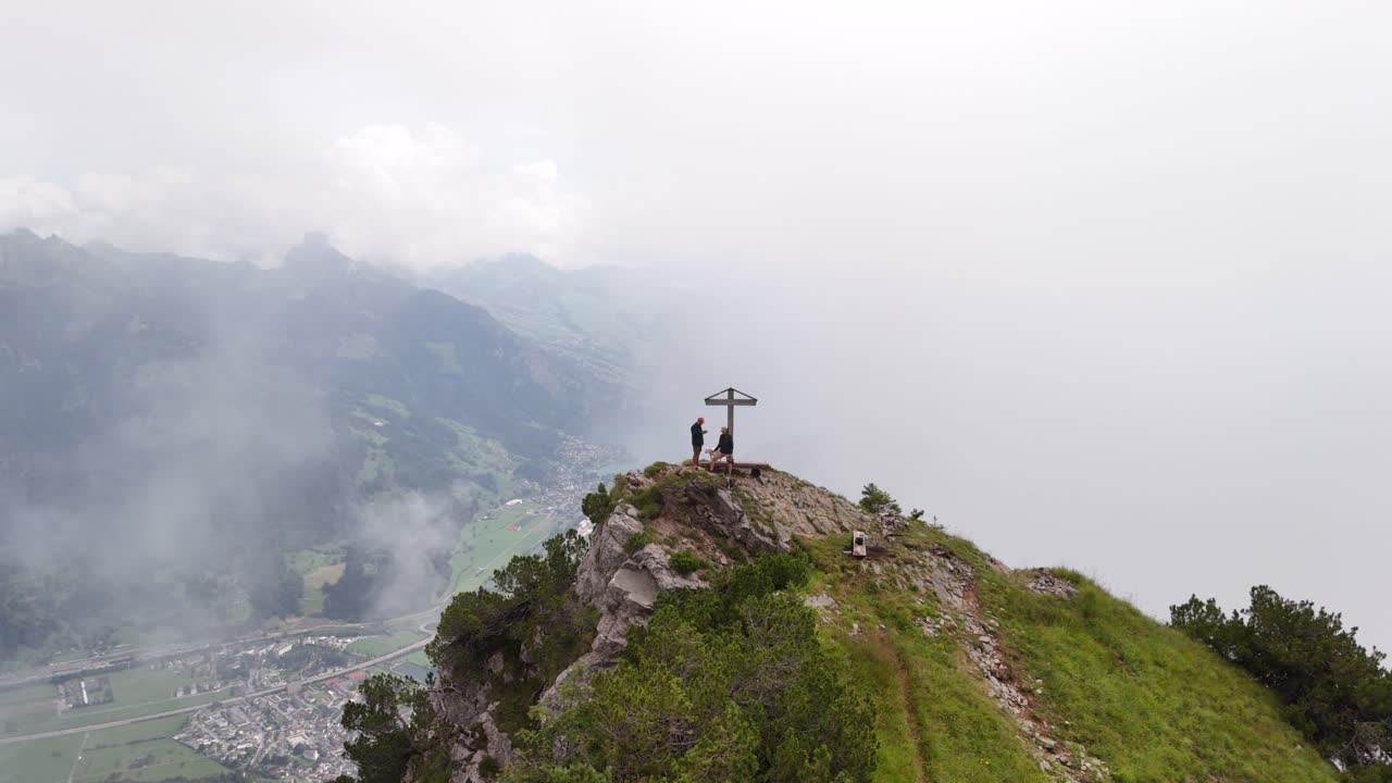 Two people standing by a cross on a mountain peak overlooking a foggy valley