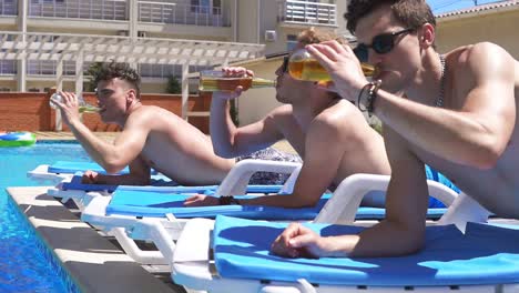 Group-of-young-friends-drinking-beer-and-relaxing-lying-on-the-coaches-by-the-swimming-pool.-Slow-Motion-shot